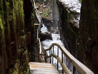 [A tall narrow opening of rock with a boardwalk and stairs against the left side and a very enthusiastic waterflow on the right side. Moss seen growing on the walls on the left.]